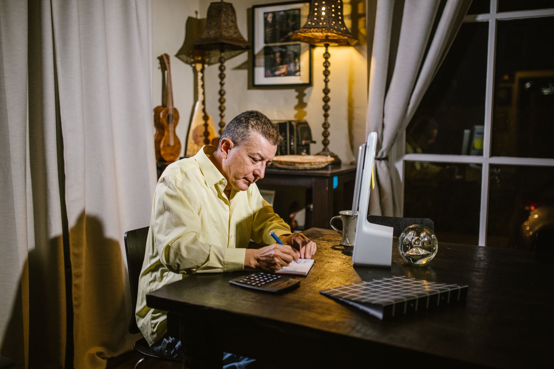 Senior man wearing yellow shirt writing notes at desk with lamp, calculator, and glass sphere nearby.
